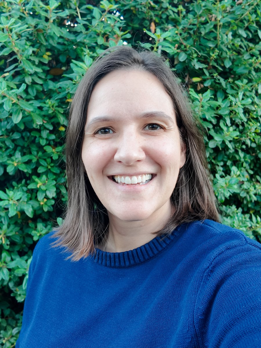 Melanie Harari smiling, in a blue sweater in front of green leaves